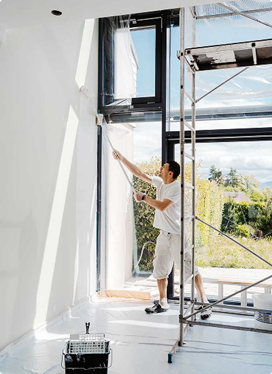 Contractor painting interior wall with roller beside scaffold by large window, Home renovation